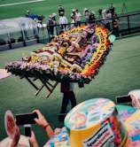 A vibrant flower arrangement carried by a person, featuring intricate patterns and text. The background includes spectators capturing the moment on their phones, and a green field with people in the distance.