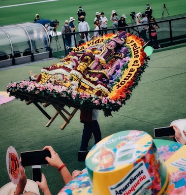 A vibrant flower arrangement carried by a person, featuring intricate patterns and text. The background includes spectators capturing the moment on their phones, and a green field with people in the distance.