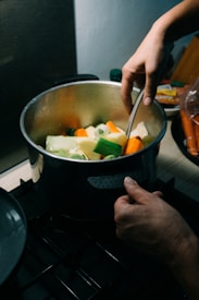 A pair of hands is stirring a pot filled with various chopped vegetables, including carrots, potatoes, and green beans. The pot sits on a stove, and the surrounding kitchen area is dimly lit, giving a warm ambiance to the scene.