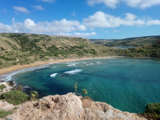 A picturesque bay with clear turquoise waters gently lapping at a sandy beach. The beach is flanked by verdant hills covered in green shrubbery. Soft, fluffy clouds float in a vibrant blue sky.