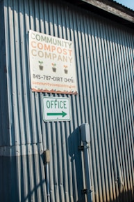 A corrugated metal wall with two signs attached. The top sign reads 'Community Compost Company' with a phone number and website. Below it, another sign indicates 'Office' with an arrow pointing right.