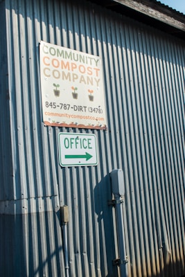 A corrugated metal wall with two signs attached. The top sign reads 'Community Compost Company' with a phone number and website. Below it, another sign indicates 'Office' with an arrow pointing right.