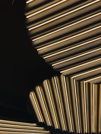 A close-up of layered, metallic surfaces featuring a ribbed pattern. The lighting and shadows create depth and a play between light and dark areas, accentuating the texture of the material.