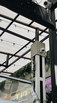 Overhead structure with metal beams and a transparent roof, decorated with string lights and green foliage. Two security cameras are mounted on a column. A partial view of glass panels and letters that spell 'EGG' in yellow can be seen.