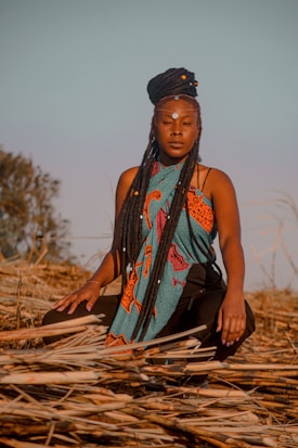 A person with long braided hair, adorned with a decorative headpiece and face paint, sits among dried reeds. Wearing a vibrant, patterned garment featuring teal and orange colors, they appear calm and introspective under a clear blue sky with soft lighting.