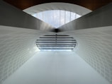 A modern architectural structure featuring a large arched ceiling with geometric patterns. The ceiling is primarily white with a subtle texture, and there is a row of windows at the top allowing natural light to illuminate the space. The design is symmetrical, creating a sense of depth and openness.