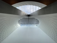 A modern architectural structure featuring a large arched ceiling with geometric patterns. The ceiling is primarily white with a subtle texture, and there is a row of windows at the top allowing natural light to illuminate the space. The design is symmetrical, creating a sense of depth and openness.
