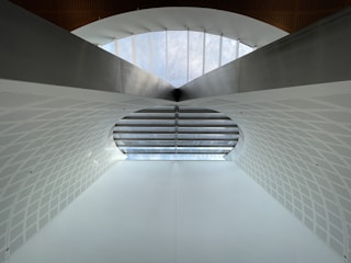A modern architectural structure featuring a large arched ceiling with geometric patterns. The ceiling is primarily white with a subtle texture, and there is a row of windows at the top allowing natural light to illuminate the space. The design is symmetrical, creating a sense of depth and openness.
