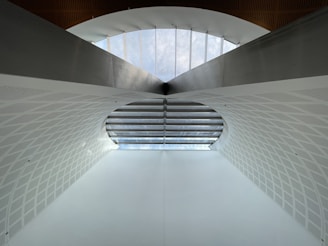 A modern architectural structure featuring a large arched ceiling with geometric patterns. The ceiling is primarily white with a subtle texture, and there is a row of windows at the top allowing natural light to illuminate the space. The design is symmetrical, creating a sense of depth and openness.