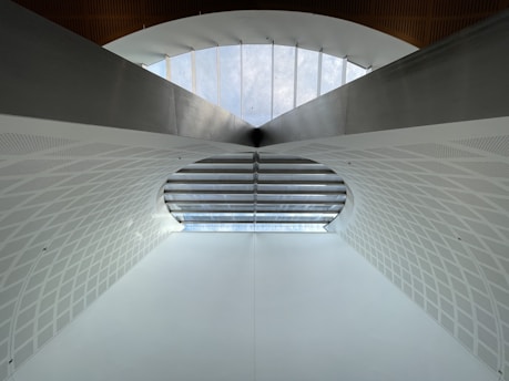A modern architectural structure featuring a large arched ceiling with geometric patterns. The ceiling is primarily white with a subtle texture, and there is a row of windows at the top allowing natural light to illuminate the space. The design is symmetrical, creating a sense of depth and openness.