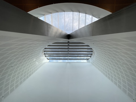 A modern architectural structure featuring a large arched ceiling with geometric patterns. The ceiling is primarily white with a subtle texture, and there is a row of windows at the top allowing natural light to illuminate the space. The design is symmetrical, creating a sense of depth and openness.