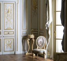 A richly decorated interior featuring ornate walls with gold detailing and intricate carvings. A classic upholstered chair with floral patterns sits on a hardwood floor, accompanied by a small decorative table. Tall windows are adorned with luxurious drapes.