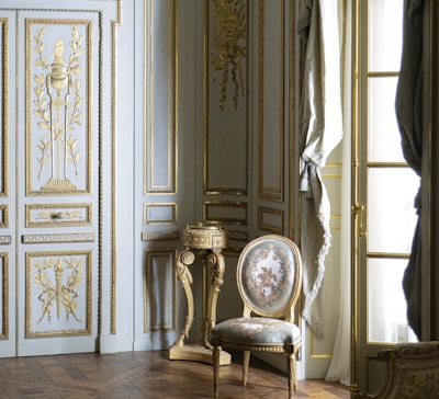 A richly decorated interior featuring ornate walls with gold detailing and intricate carvings. A classic upholstered chair with floral patterns sits on a hardwood floor, accompanied by a small decorative table. Tall windows are adorned with luxurious drapes.