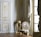 A richly decorated interior featuring ornate walls with gold detailing and intricate carvings. A classic upholstered chair with floral patterns sits on a hardwood floor, accompanied by a small decorative table. Tall windows are adorned with luxurious drapes.