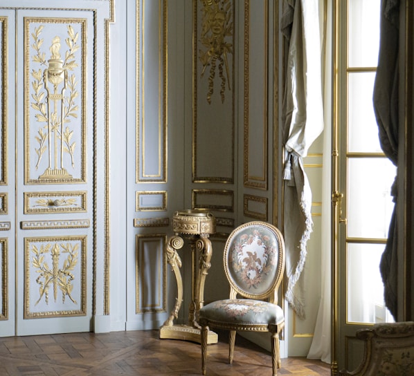 A richly decorated interior featuring ornate walls with gold detailing and intricate carvings. A classic upholstered chair with floral patterns sits on a hardwood floor, accompanied by a small decorative table. Tall windows are adorned with luxurious drapes.