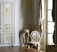 A richly decorated interior featuring ornate walls with gold detailing and intricate carvings. A classic upholstered chair with floral patterns sits on a hardwood floor, accompanied by a small decorative table. Tall windows are adorned with luxurious drapes.