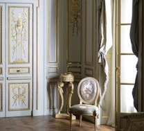 A richly decorated interior featuring ornate walls with gold detailing and intricate carvings. A classic upholstered chair with floral patterns sits on a hardwood floor, accompanied by a small decorative table. Tall windows are adorned with luxurious drapes.
