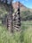 A rustic wooden fence with a distinctive radial design stands among tall grass in a natural landscape. In the background, rocky hills rise against a partly cloudy sky, creating an earthy and serene atmosphere. The surrounding greenery consists of trees and shrubs, adding to the tranquility of the scene.