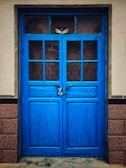 A vibrant blue, double-panel wooden door with glass panes and a visible padlock in the center. The door is set within a beige wall and bordered by two textured brick columns.
