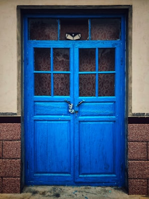 A vibrant blue, double-panel wooden door with glass panes and a visible padlock in the center. The door is set within a beige wall and bordered by two textured brick columns.