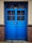 A vibrant blue, double-panel wooden door with glass panes and a visible padlock in the center. The door is set within a beige wall and bordered by two textured brick columns.