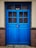 A vibrant blue, double-panel wooden door with glass panes and a visible padlock in the center. The door is set within a beige wall and bordered by two textured brick columns.