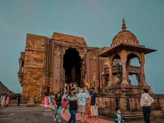 A historic stone temple with intricate carvings and architecture is depicted. People are walking around the temple, and some are wearing face masks, suggesting a contemporary setting. The sky is clear, providing a serene backdrop to the ancient structure.