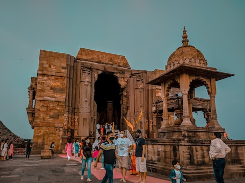 A historic stone temple with intricate carvings and architecture is depicted. People are walking around the temple, and some are wearing face masks, suggesting a contemporary setting. The sky is clear, providing a serene backdrop to the ancient structure.
