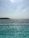 A swimming pool with several swimmers is situated alongside a vast body of water, possibly the ocean. The sunlight reflects on the pool, creating a shimmering effect. In the background, the horizon stretches with a hazy sky and a distant coastline.