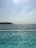 A swimming pool with several swimmers is situated alongside a vast body of water, possibly the ocean. The sunlight reflects on the pool, creating a shimmering effect. In the background, the horizon stretches with a hazy sky and a distant coastline.