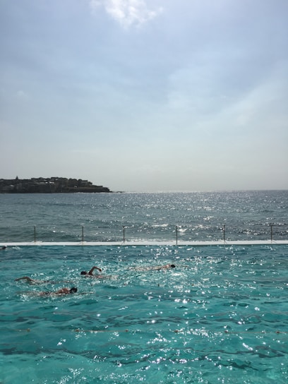 A swimming pool with several swimmers is situated alongside a vast body of water, possibly the ocean. The sunlight reflects on the pool, creating a shimmering effect. In the background, the horizon stretches with a hazy sky and a distant coastline.