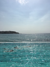 A swimming pool with several swimmers is situated alongside a vast body of water, possibly the ocean. The sunlight reflects on the pool, creating a shimmering effect. In the background, the horizon stretches with a hazy sky and a distant coastline.