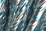 A close-up view of twisted, metallic surfaces with a smooth, reflective texture. The surfaces have an intricate pattern, featuring shiny blue and silver hues that create a futuristic and abstract visual effect.