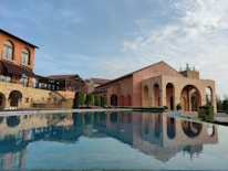 A scenic view of a Mediterranean-style villa with warm-toned walls, featuring arched windows and doorways. In the foreground, there is a reflective swimming pool that mirrors the building's architecture, surrounded by neatly trimmed bushes and outdoor seating areas. The sky is partly cloudy, adding to the serene ambiance of the scene.