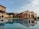 A scenic view of a Mediterranean-style villa with warm-toned walls, featuring arched windows and doorways. In the foreground, there is a reflective swimming pool that mirrors the building's architecture, surrounded by neatly trimmed bushes and outdoor seating areas. The sky is partly cloudy, adding to the serene ambiance of the scene.