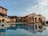 A scenic view of a Mediterranean-style villa with warm-toned walls, featuring arched windows and doorways. In the foreground, there is a reflective swimming pool that mirrors the building's architecture, surrounded by neatly trimmed bushes and outdoor seating areas. The sky is partly cloudy, adding to the serene ambiance of the scene.
