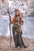 A woman dressed in a fantasy or medieval-style costume stands by a serene lake. She holds a large, intricately designed staff and wears detailed armor and accessories. Her long, red hair and the overall earthy tones of her outfit contrast with the natural, muted background of trees and water.