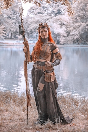 A woman dressed in a fantasy or medieval-style costume stands by a serene lake. She holds a large, intricately designed staff and wears detailed armor and accessories. Her long, red hair and the overall earthy tones of her outfit contrast with the natural, muted background of trees and water.