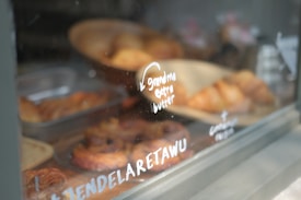 A display window containing various pastries, including croissants and other baked goods. The glass has writing on it, including phrases like 'Grandma extra butter' in white lettering. The pastries are arranged in metal trays and a wooden basket, creating an inviting atmosphere.
