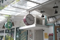 A shop facade with hanging signs, including a round sign for 'Suwe Ora Jamu'. Vintage-style hanging lamps are attached to the ceiling. There is visible greenery, a glass canopy, and an air conditioning unit.