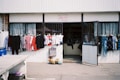 A small shop with an open entrance displaying a variety of clothing items outside. Brightly colored shirts and dresses hang on racks, while other clothes are visible through the shop's doorway. The shop front is simple, with a sign above the door and a concrete sidewalk in front.