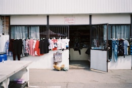 A small shop with an open entrance displaying a variety of clothing items outside. Brightly colored shirts and dresses hang on racks, while other clothes are visible through the shop's doorway. The shop front is simple, with a sign above the door and a concrete sidewalk in front.