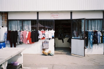 A small shop with an open entrance displaying a variety of clothing items outside. Brightly colored shirts and dresses hang on racks, while other clothes are visible through the shop's doorway. The shop front is simple, with a sign above the door and a concrete sidewalk in front.