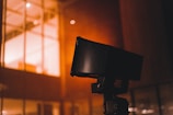 A close-up of a black security camera mounted on a structure, with a bright, warmly lit building with windows in the background. Some raindrops or moisture appear to be present on the camera, suggesting a recent rainfall or high humidity.