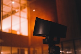 A close-up of a black security camera mounted on a structure, with a bright, warmly lit building with windows in the background. Some raindrops or moisture appear to be present on the camera, suggesting a recent rainfall or high humidity.