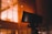 A close-up of a black security camera mounted on a structure, with a bright, warmly lit building with windows in the background. Some raindrops or moisture appear to be present on the camera, suggesting a recent rainfall or high humidity.