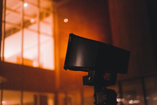 A close-up of a black security camera mounted on a structure, with a bright, warmly lit building with windows in the background. Some raindrops or moisture appear to be present on the camera, suggesting a recent rainfall or high humidity.