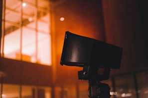 A close-up of a black security camera mounted on a structure, with a bright, warmly lit building with windows in the background. Some raindrops or moisture appear to be present on the camera, suggesting a recent rainfall or high humidity.