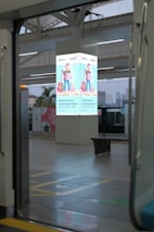 A modern train station platform is visible, featuring a lit advertisement on a central pillar. The ad displays a man holding shopping bags and wearing a casual outfit with a guitar next to him. Surrounding the platform is an urban environment with skyscrapers in the background, and some greenery is visible. The scene is observed through the open door of a train.