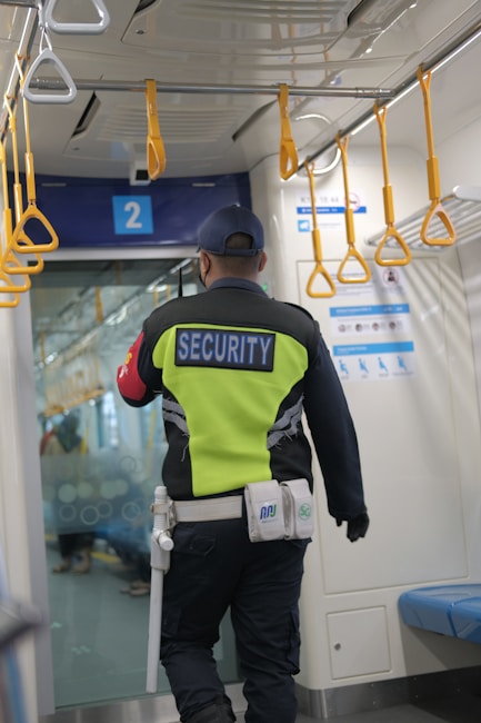 A security guard with a bright yellow vest and the word 'SECURITY' on the back stands inside a public transportation vehicle. Overhead are several orange hand grips for passengers. The seating area features blue seats, and there are informational signs and a blue door marked with the number '2'.
